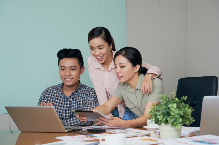 Three people collaborating around a laptop and tablet at a table with papers and a plant.