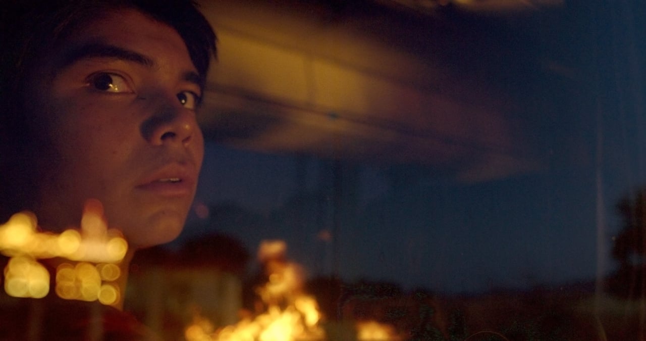 A person with a serious expression gazes out of a window at night, with reflections of lights and blurred shapes visible.
