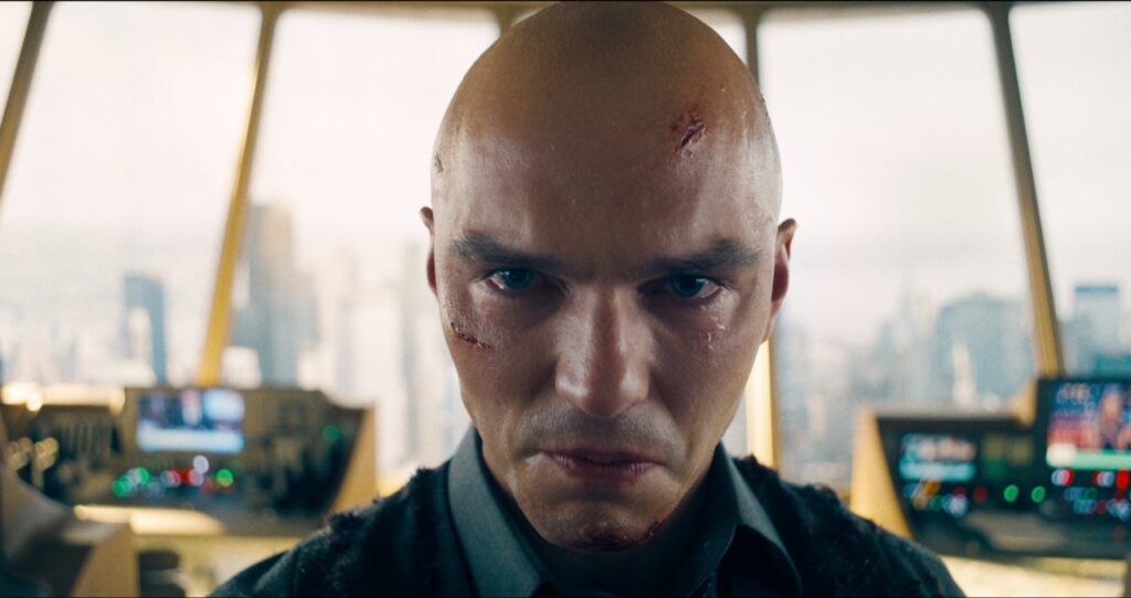 A bald man with facial scars is standing in a control room with blurred cityscape visible through the windows.
