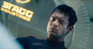 Everything We Know About James Gunn’s Superman Movie 2025 A man in a dark suit with eyes closed, standing under a sign that says "Stagg Industries.
