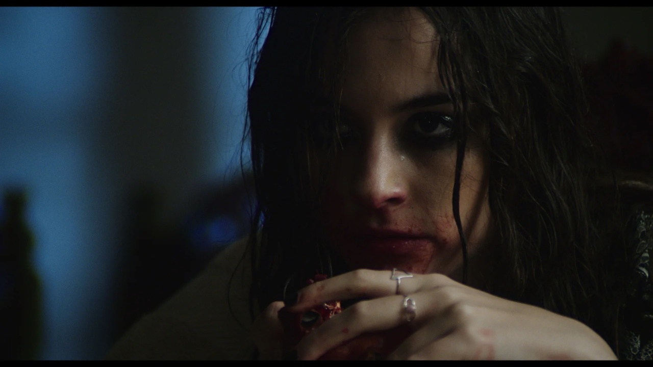 A person with wet hair, dark eye makeup, and stained lips is holding an object near their face. The setting is dimly lit.