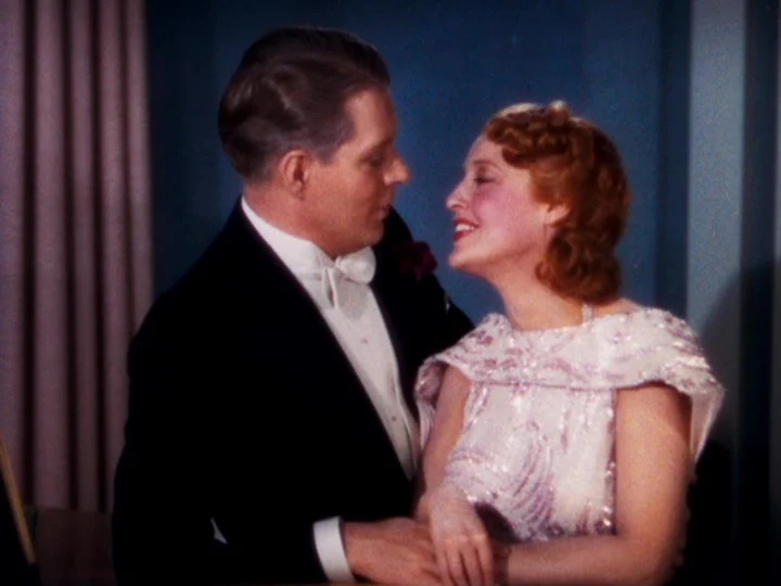A couple in formal attire, with the man in a tuxedo and the woman in a sparkling gown, share a moment of laughter against a blue background.