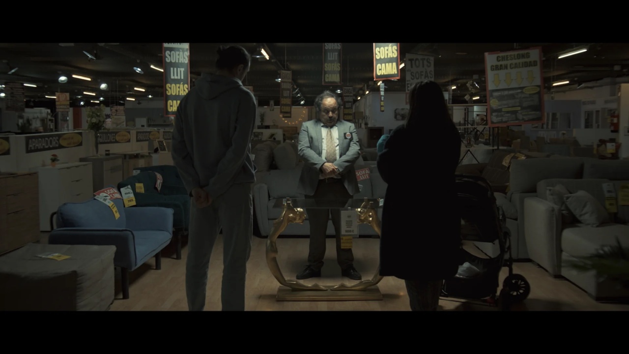 Two people stand in a dimly lit furniture store, observing a man in a suit standing behind a golden mirror. Various signs and furniture pieces are visible in the background.