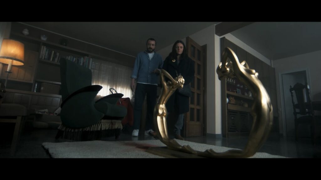 A man and woman stand in a dimly lit room, looking at an ornate, golden, double-headed snake sculpture on the floor.