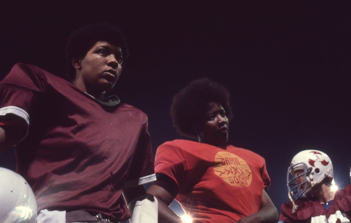 Football players stand together on a field under stadium lights, wearing maroon and red jerseys, with helmets held in their hands.