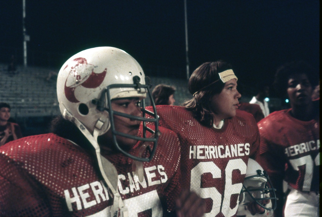 Football players wearing red jerseys with "HERRICANES" on the front stand on the sideline. One player wears a helmet, while another has a headband. The field lights are on.