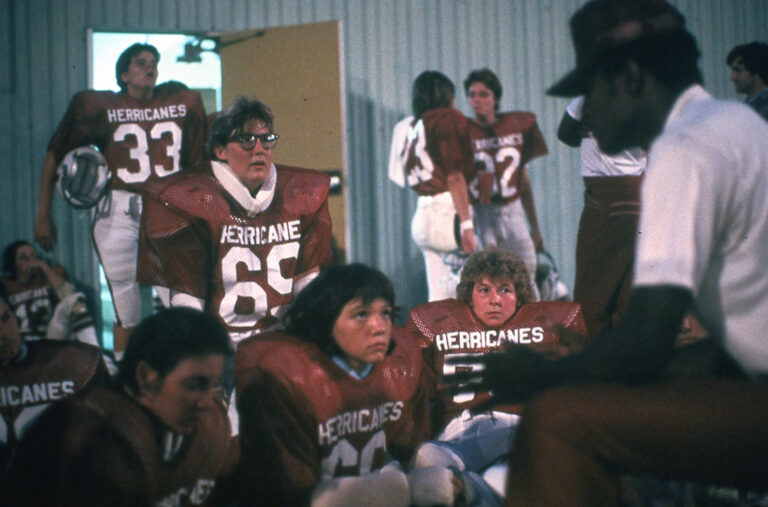 Football players in red jerseys sit and listen to a coach indoors. Jerseys read "Hurricanes." One player with glasses is number 69.