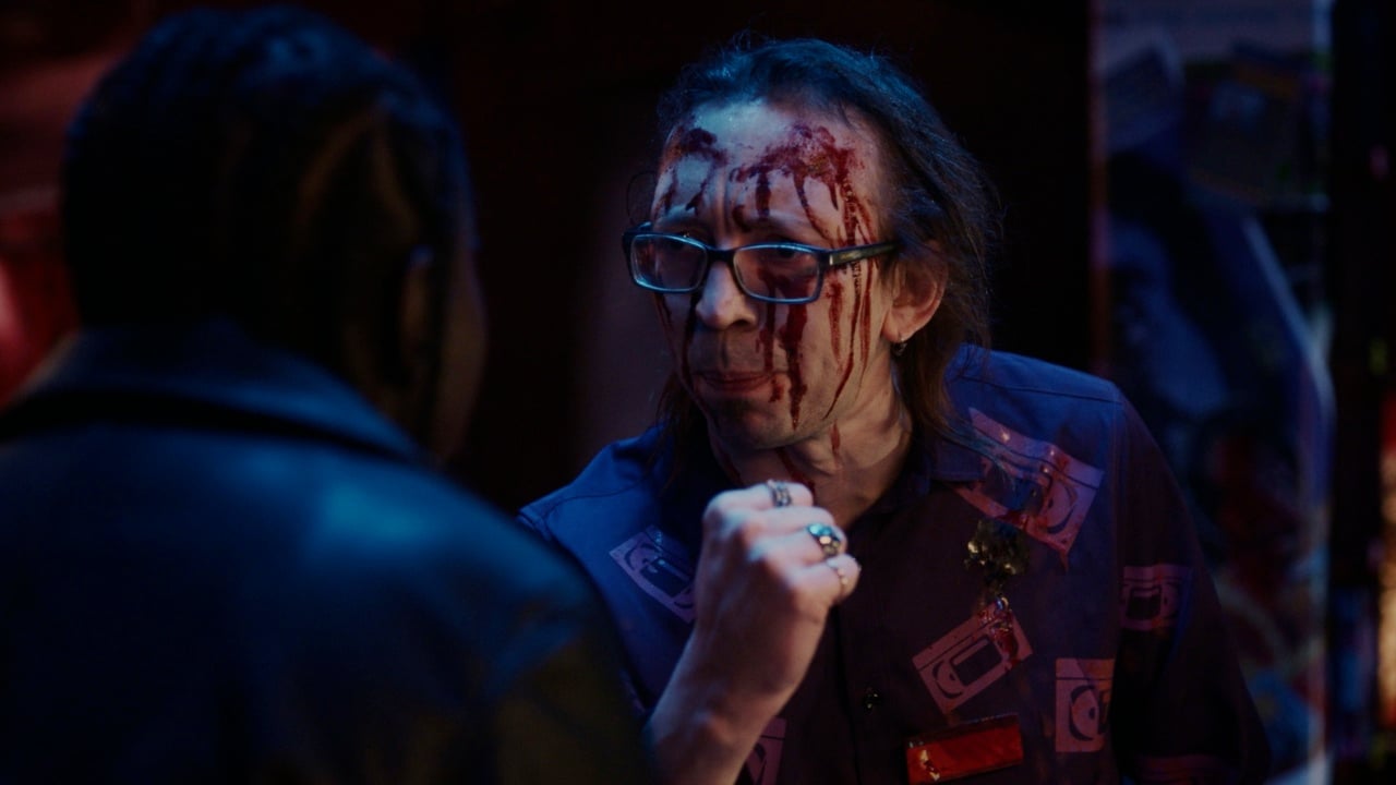 A man wearing glasses and a patterned shirt with blood on his face gestures toward a person in the foreground.