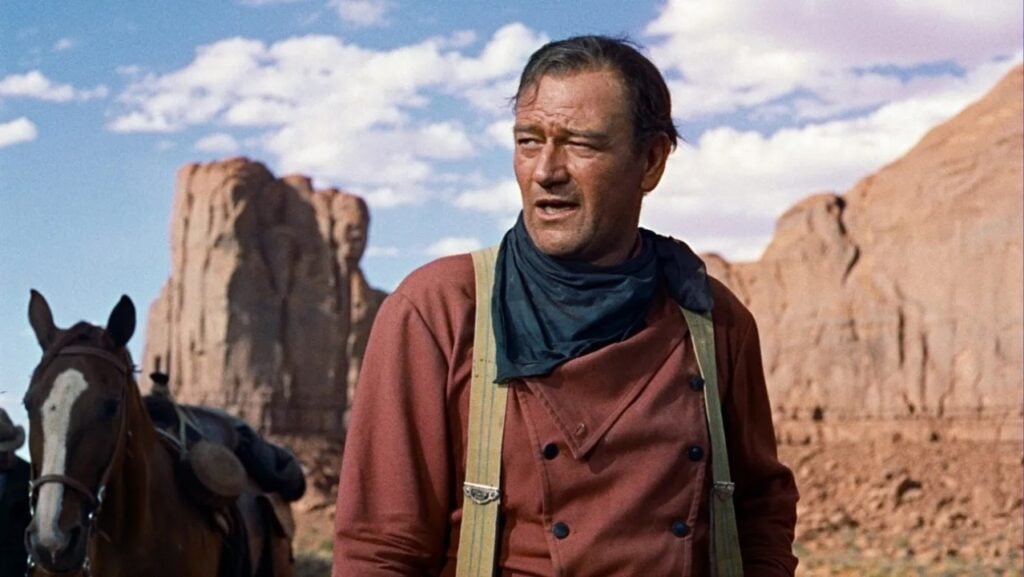 A person in Western attire stands outdoors in a desert landscape with rock formations and a horse in the background.