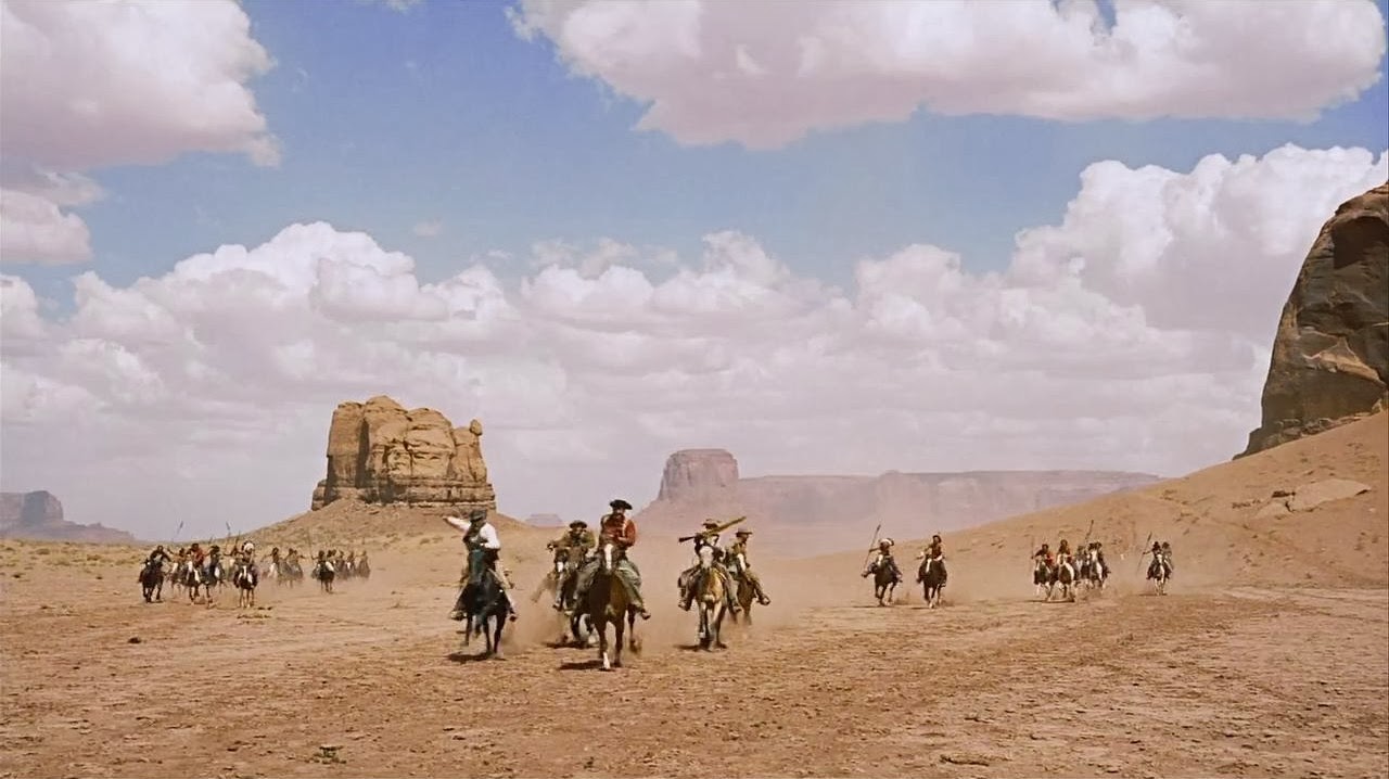 A group of people on horseback rides across a desert landscape under a partly cloudy sky..