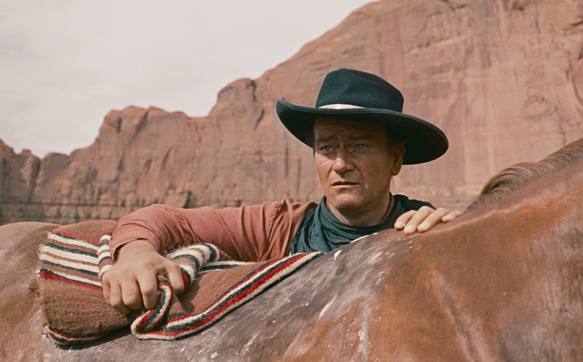 A person wearing a black hat leans on a horse, set against a backdrop of red rock formations.