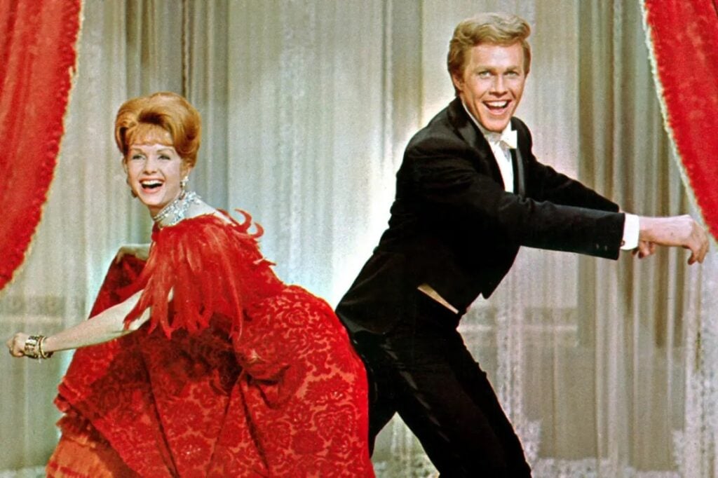 Two people in formal attire perform a playful dance routine, smiling widely. The woman wears a red dress, and the man is in a tuxedo.