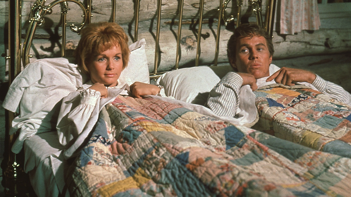 A man and woman lie in bed under patchwork quilts, both looking towards the same direction.