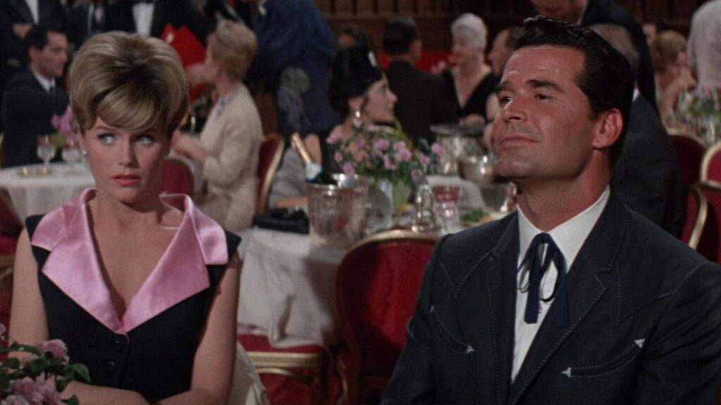 A woman and a man sit at a formal event with tables and people in the background. The woman wears a black dress with a pink collar, and the man wears a dark suit with a bolo tie.
