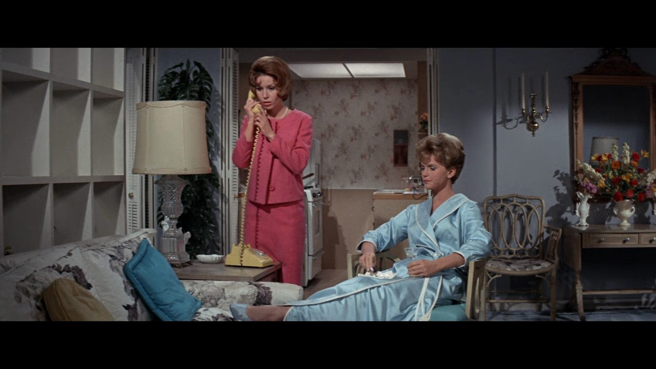 Two women in a living room; one is standing and talking on the phone, wearing a pink suit. The other is sitting on a couch, wearing a blue robe and holding a tissue.