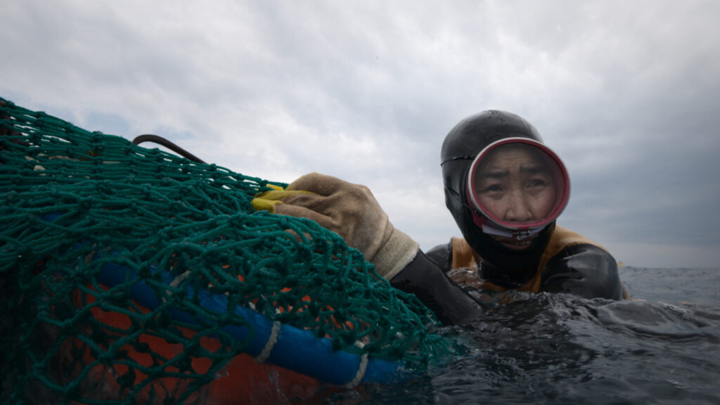 A person in a wetsuit and goggles holds onto a large net while floating in the sea under a cloudy sky.
