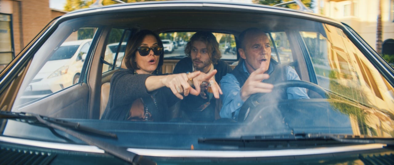 Three people are sitting in a car, looking forward. The woman on the left points ahead, while the man on the right gestures while driving. The man in the middle looks in the same direction.