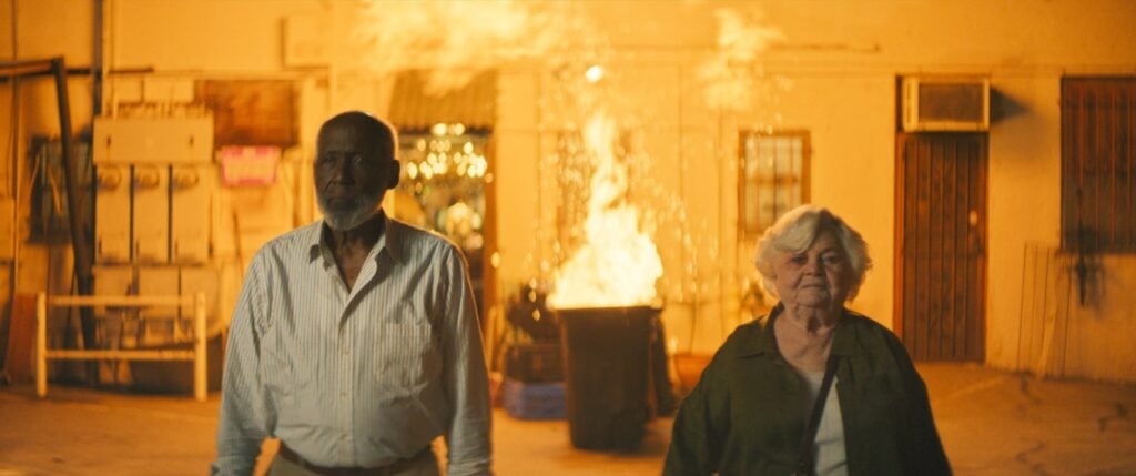 An older man and woman walk sternly toward the camera, with a large fire burning in a barrel behind them.