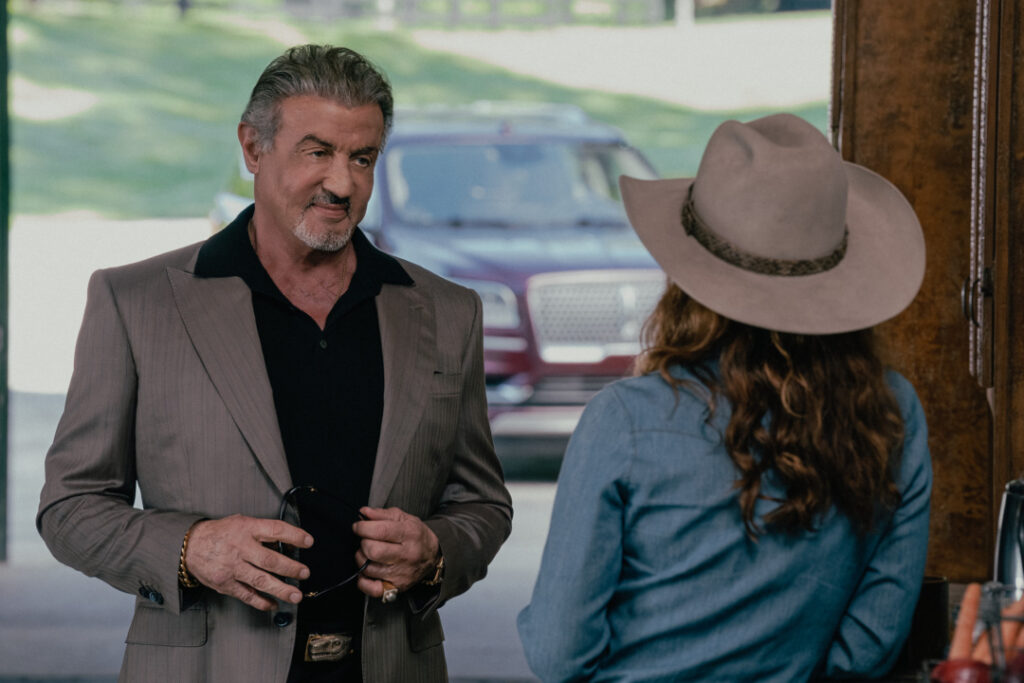 A man in a suit talks to a woman wearing a cowboy hat in an outdoor setting. A vehicle is parked in the background.
