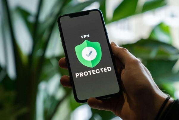 A hand holds a smartphone displaying a "VPN Protected" message with a green shield icon, against a blurred background with green foliage.