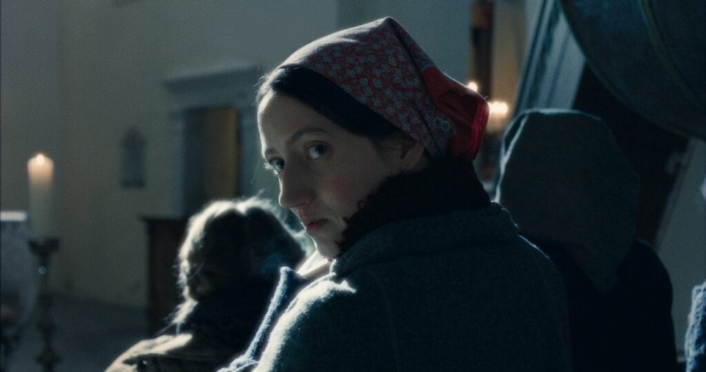 A woman in a headscarf looks over her shoulder in a dimly lit room with candles in the background.