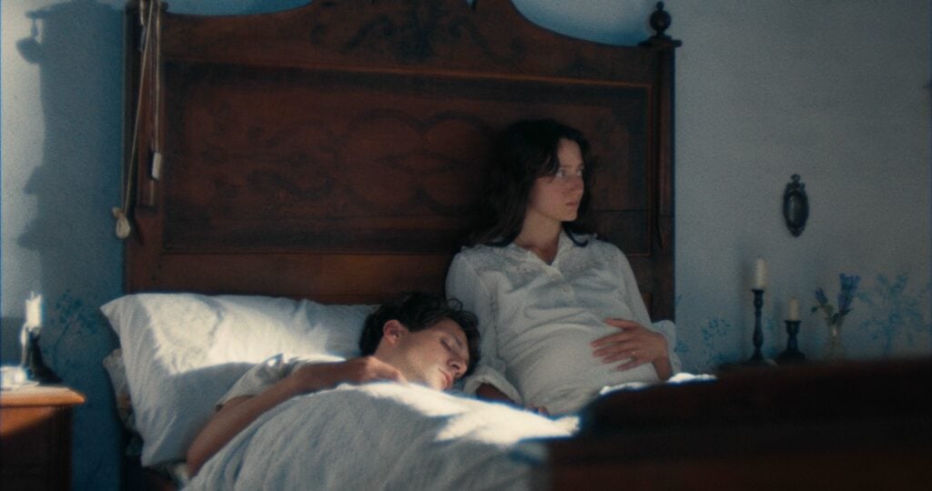 A woman sits on a bed, looking away, with her hand on her belly. A person is lying next to her, resting on a pillow. The room has a wooden headboard and simple decor.