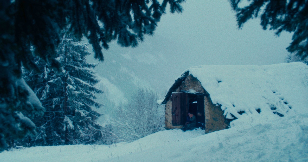 A small, snow-covered cabin in a winter landscape, surrounded by trees. A person is seated at the entrance, partially visible.