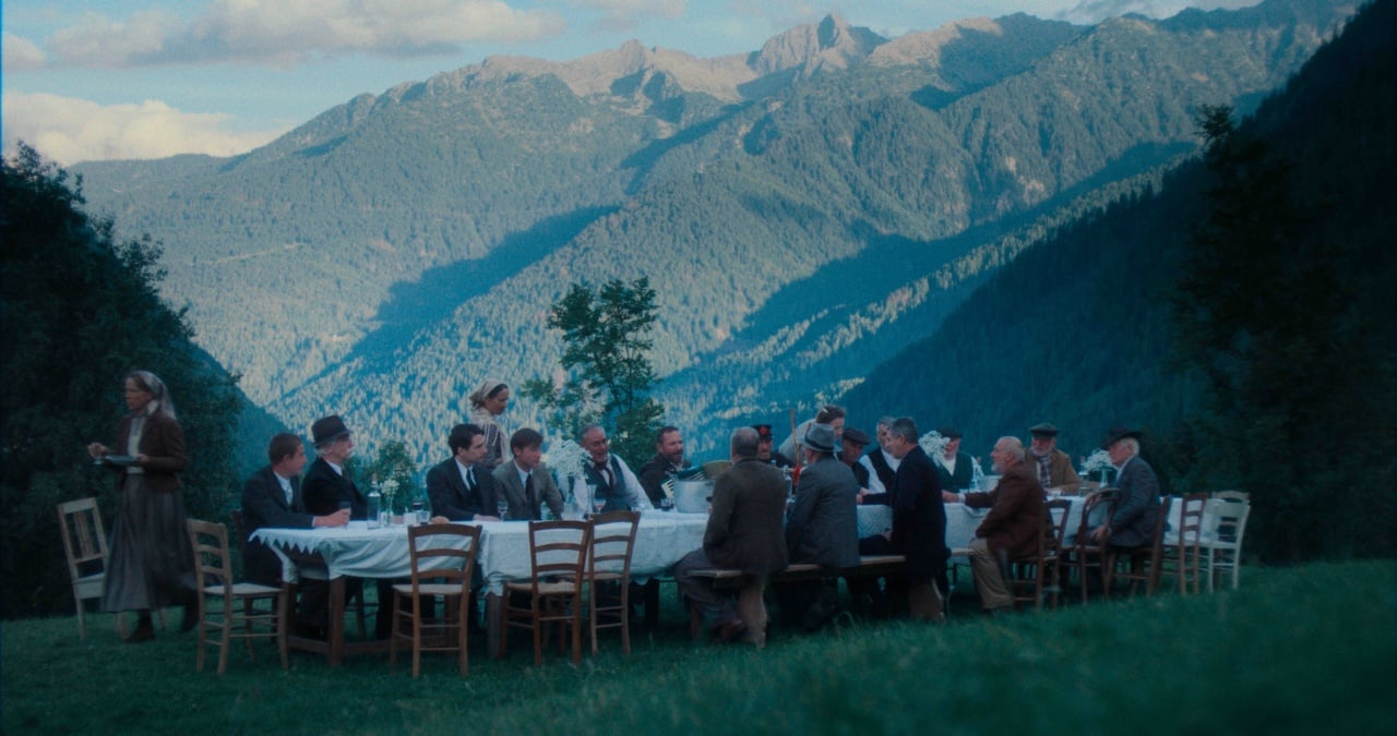 A group of people sit around a long table outdoors, dining on a grassy area with mountains in the background.