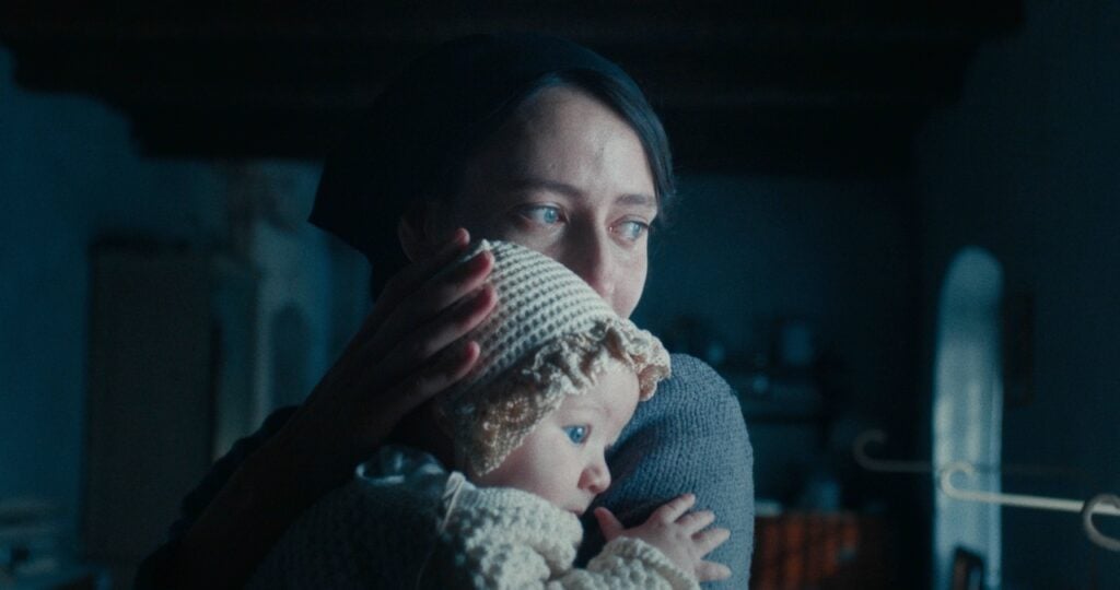 A woman in a dark headscarf holds a baby wearing a crocheted bonnet, gazing into the distance in a dimly lit room.