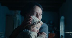 A woman in a dark headscarf holds a baby wearing a crocheted bonnet, gazing into the distance in a dimly lit room.