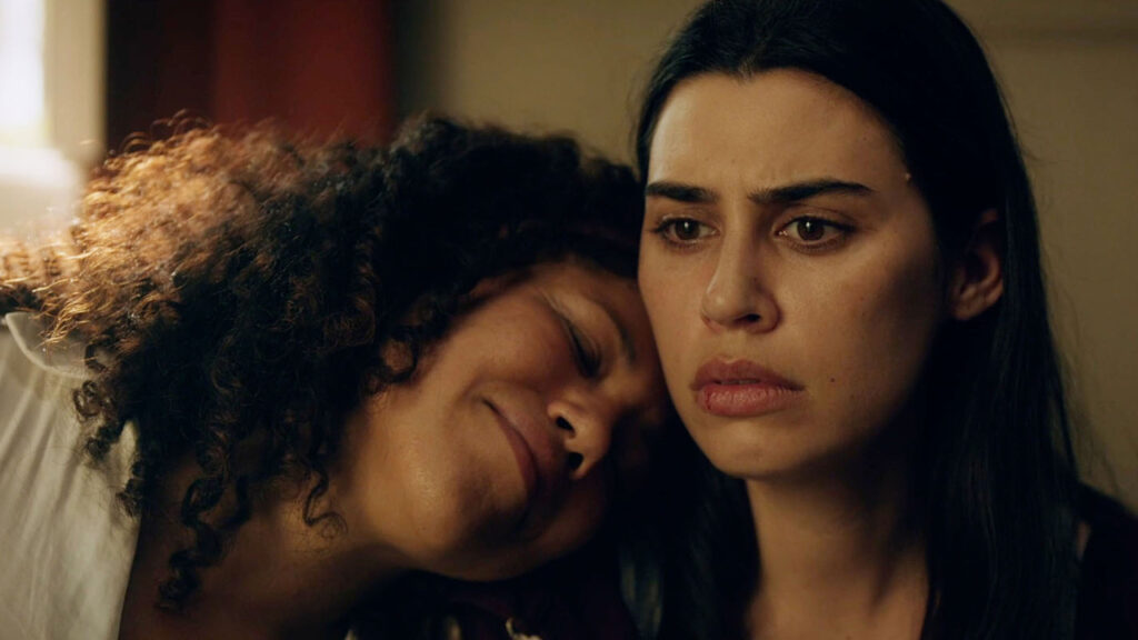 Two women sit closely; one with curly hair rests her head on the other's shoulder, who has straight black hair and a serious expression.