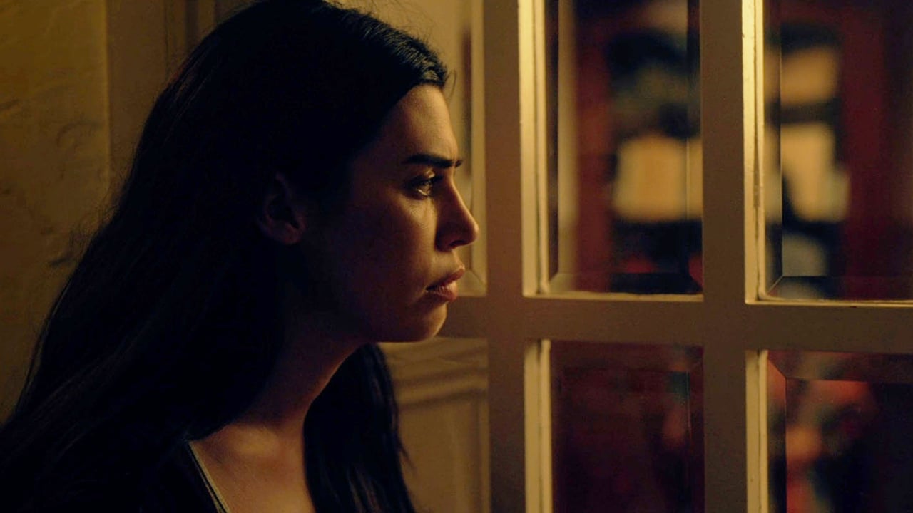 Woman with long dark hair looking out through a window, light illuminating her face against a dim background.