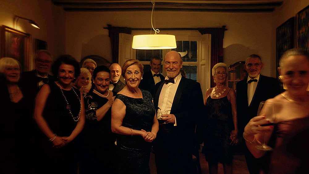 A group of elegantly dressed people poses in a dimly lit room, with a couple in the center holding drinks and smiling at the camera.
