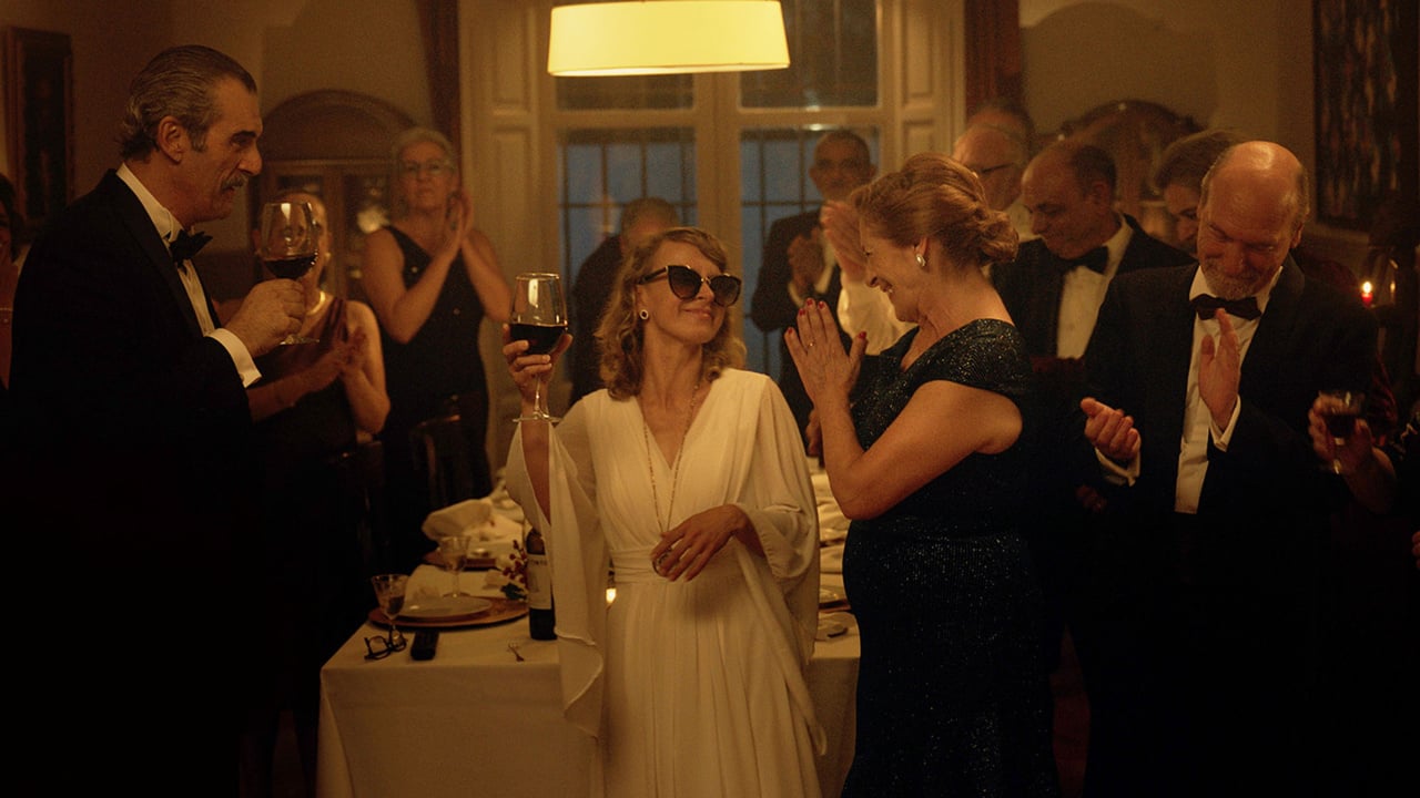 A woman in a white dress and sunglasses raises a wine glass in a dimly lit room, surrounded by elegantly dressed people applauding.