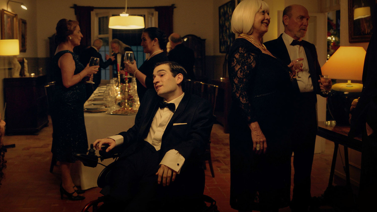 A man in a tuxedo sits in a wheelchair at a formal gathering, surrounded by people in evening attire holding drinks.