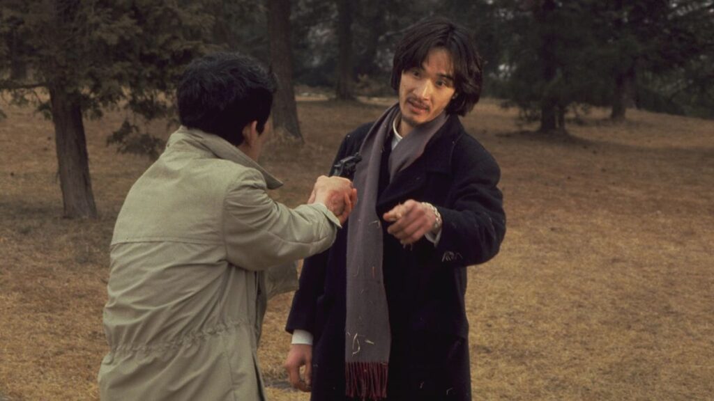 Two men in a park, one pointing a gun at the other. Trees and fallen leaves in the background.