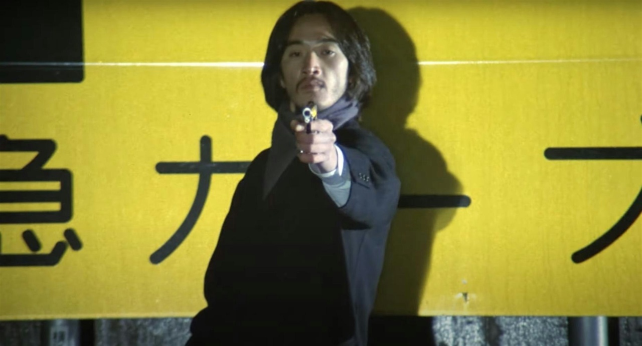 A person wearing a scarf points a gun directly at the camera, standing against a yellow sign with Japanese characters.