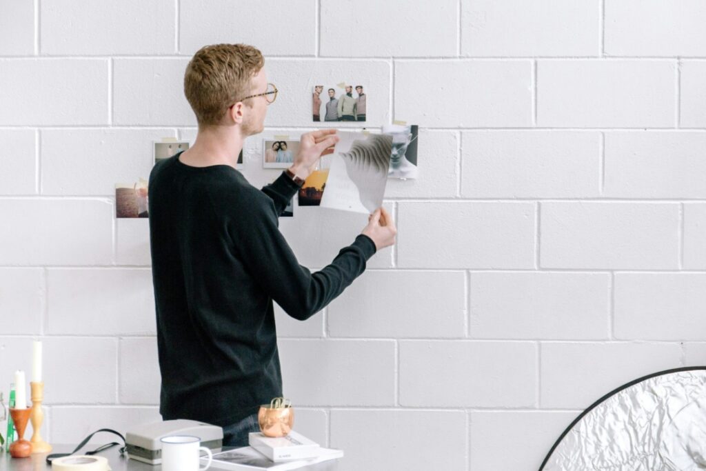 Person in black sweater arranging photos on a white brick wall, standing beside a table with various items, including a candle and a cup.