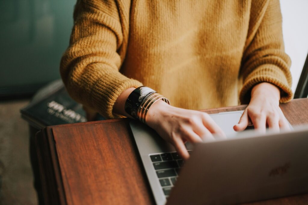 A person in a yellow sweater types on a laptop at a wooden desk.