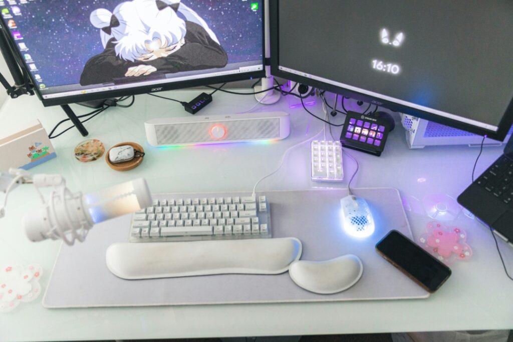 Dual-monitor computer setup with a white keyboard, a glowing mouse, a microphone, and a smartphone on a desk. Monitors display an anime character and the time 16:10.