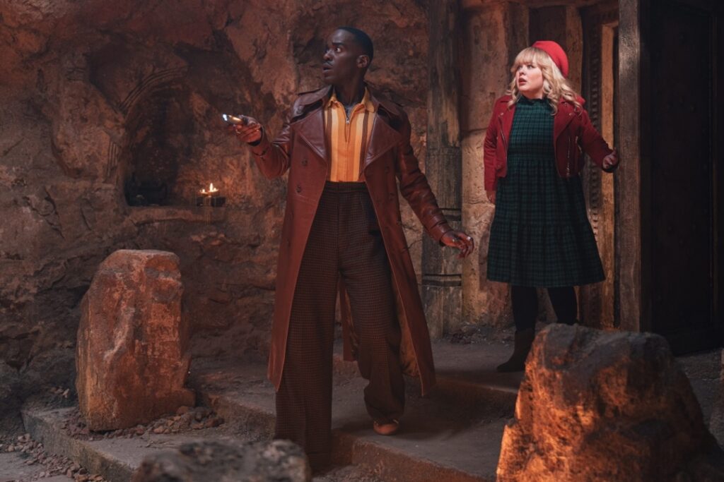 Two people stand in a dimly lit, rocky cave. One holds a device, and both appear alert. There are stone pillars and a small lit candle nearby.