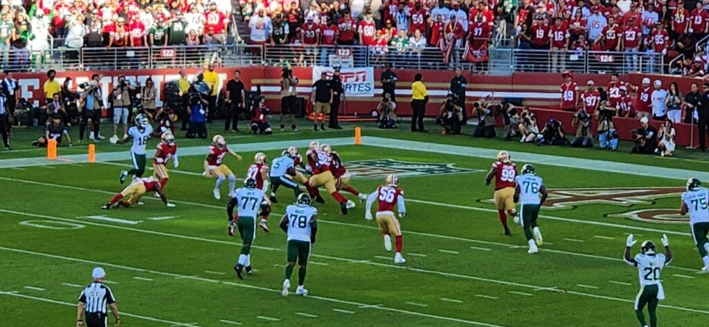Football players in red and white uniforms, with opposing team in green and white, in the middle of a play on a green field. Stadium crowd in the background.