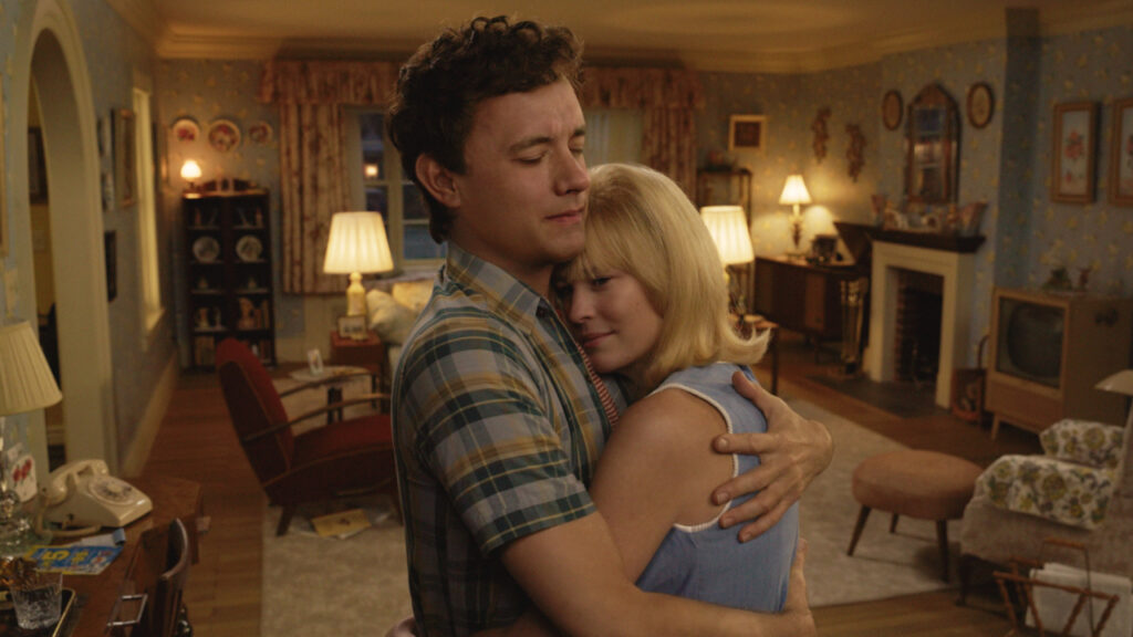 A man and woman embrace in a cozy, vintage living room with warm lighting. The room features a patterned wallpaper, bookshelves, lamps, and a television.