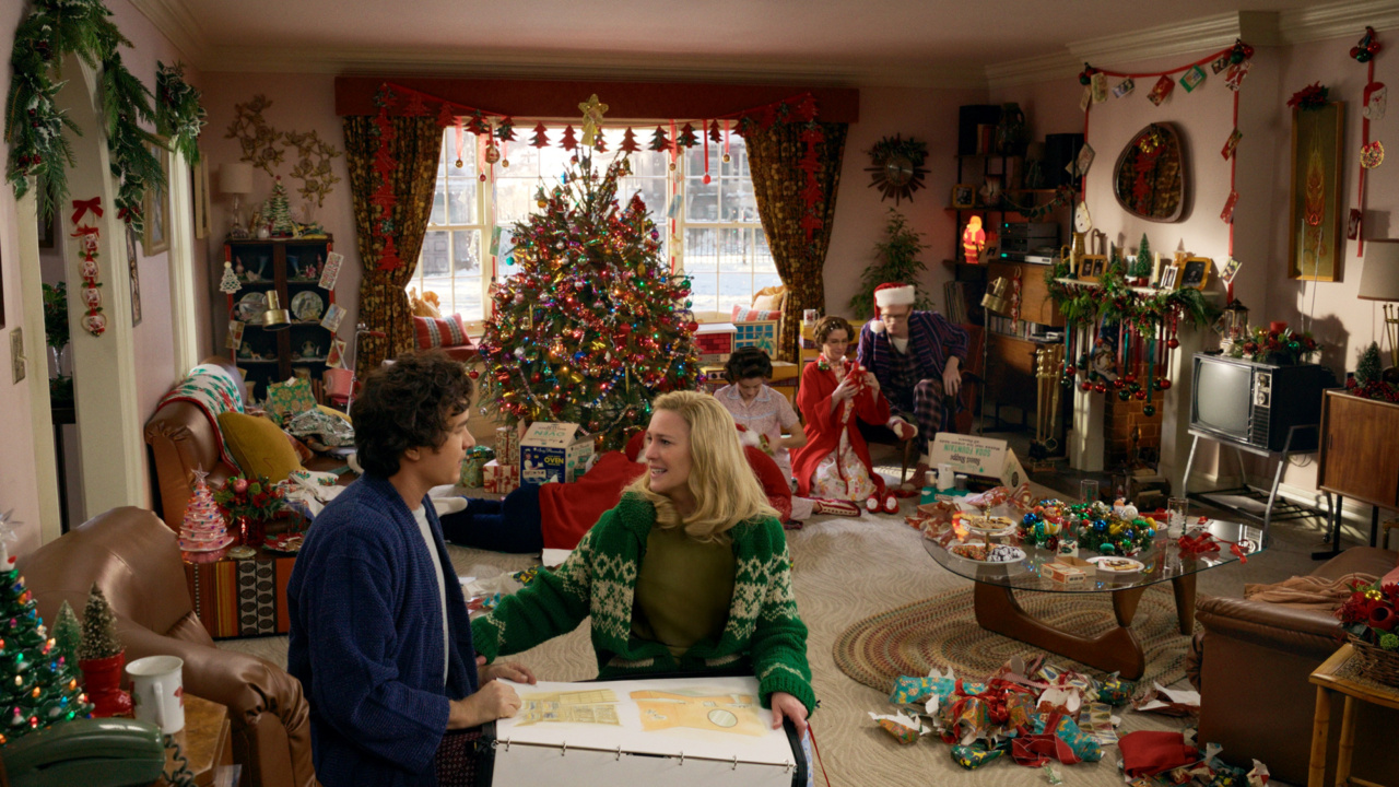 Family in a festively decorated living room unwraps gifts around a Christmas tree, surrounded by holiday decorations and wrapping paper.