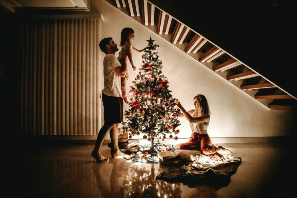 A family decorates a lit Christmas tree under a staircase. The father lifts a young girl to place an ornament, while the mother arranges decorations on the lower branches.