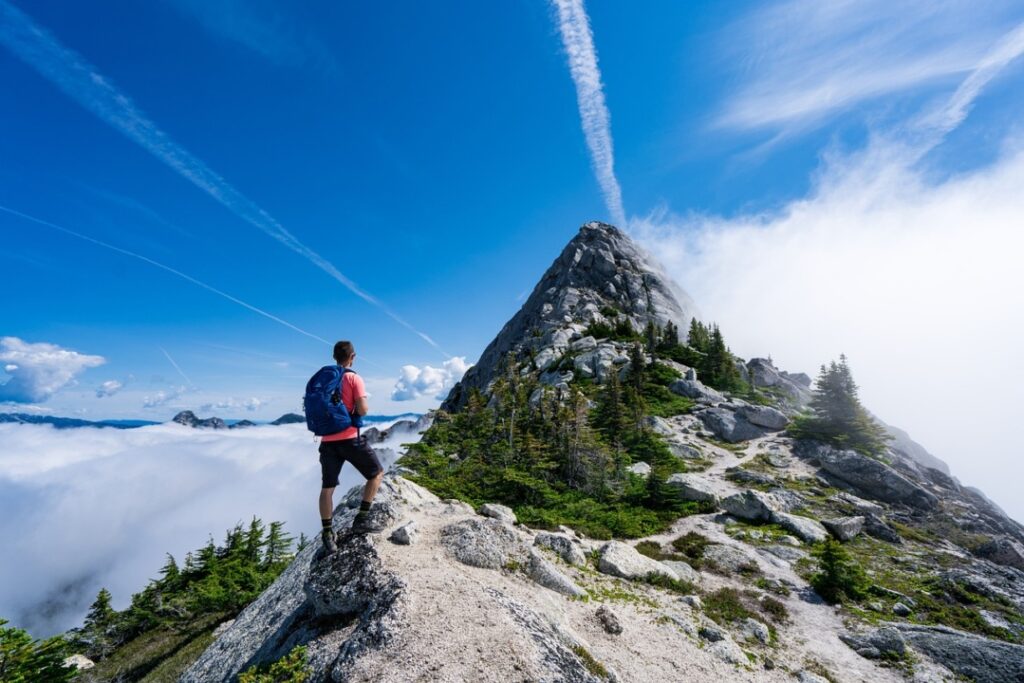A person with a backpack stands on a rocky mountain path, admiring the peak ahead. Trails of clouds and jet contrails stretch across the blue sky.