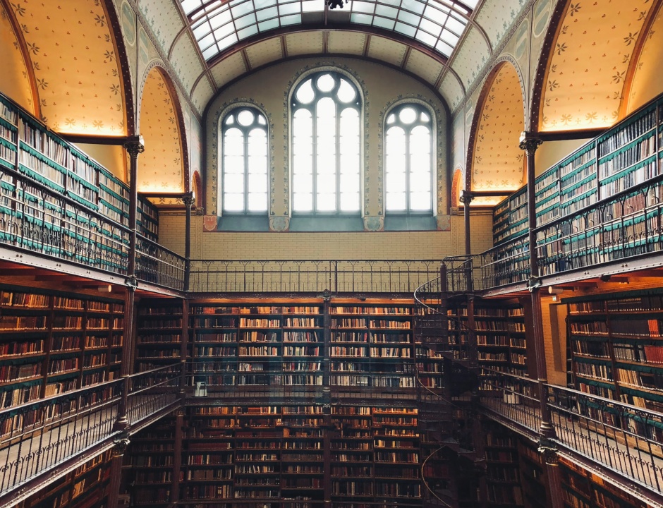 Interior of a large, ornate library with two floors of bookshelves, arched windows, and ornate metalwork railings. A spiral staircase connects the levels.