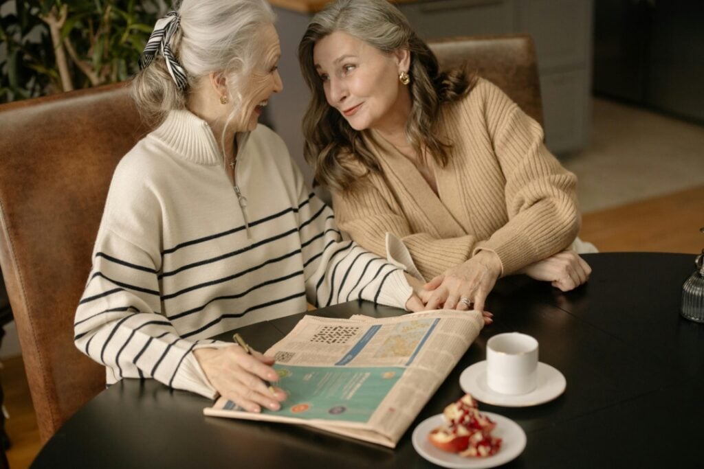 Two women sit at a table with a newspaper and dessert. They are smiling at each other, with one holding a pen and the other resting her hand on the newspaper.