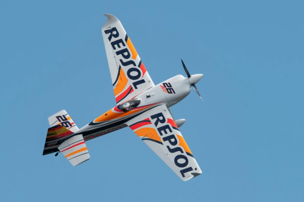 Aerobatic airplane with "Repsol" branding performs a maneuver against a clear blue sky.