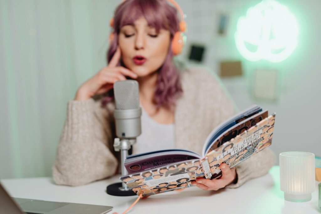 Person with purple hair wearing headphones, speaking into a microphone while holding an open book. Neon sign in the background.